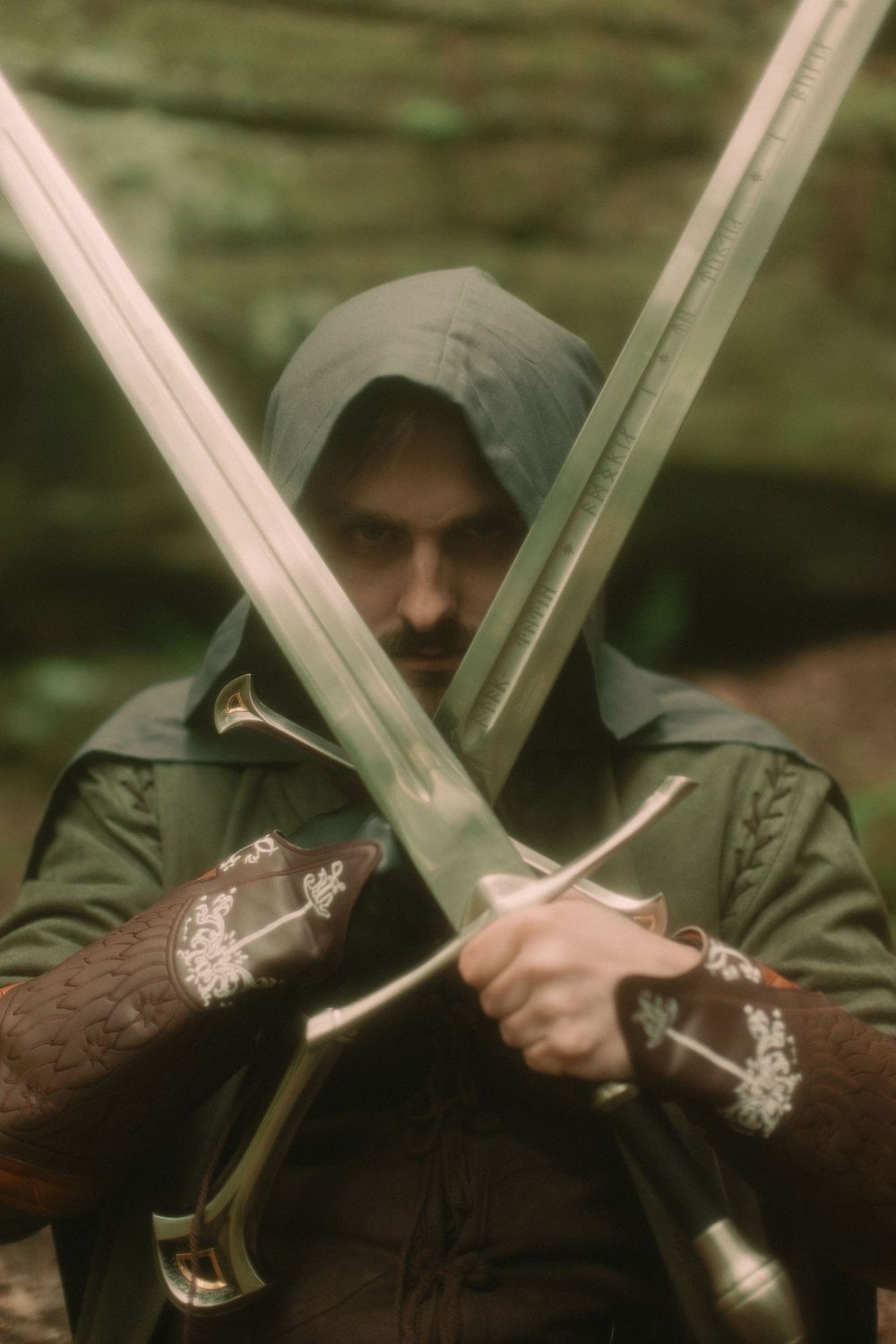 Man in a mossy forest wearing a cloak and warrior attire holding two swords in front of his face while making a serious expression