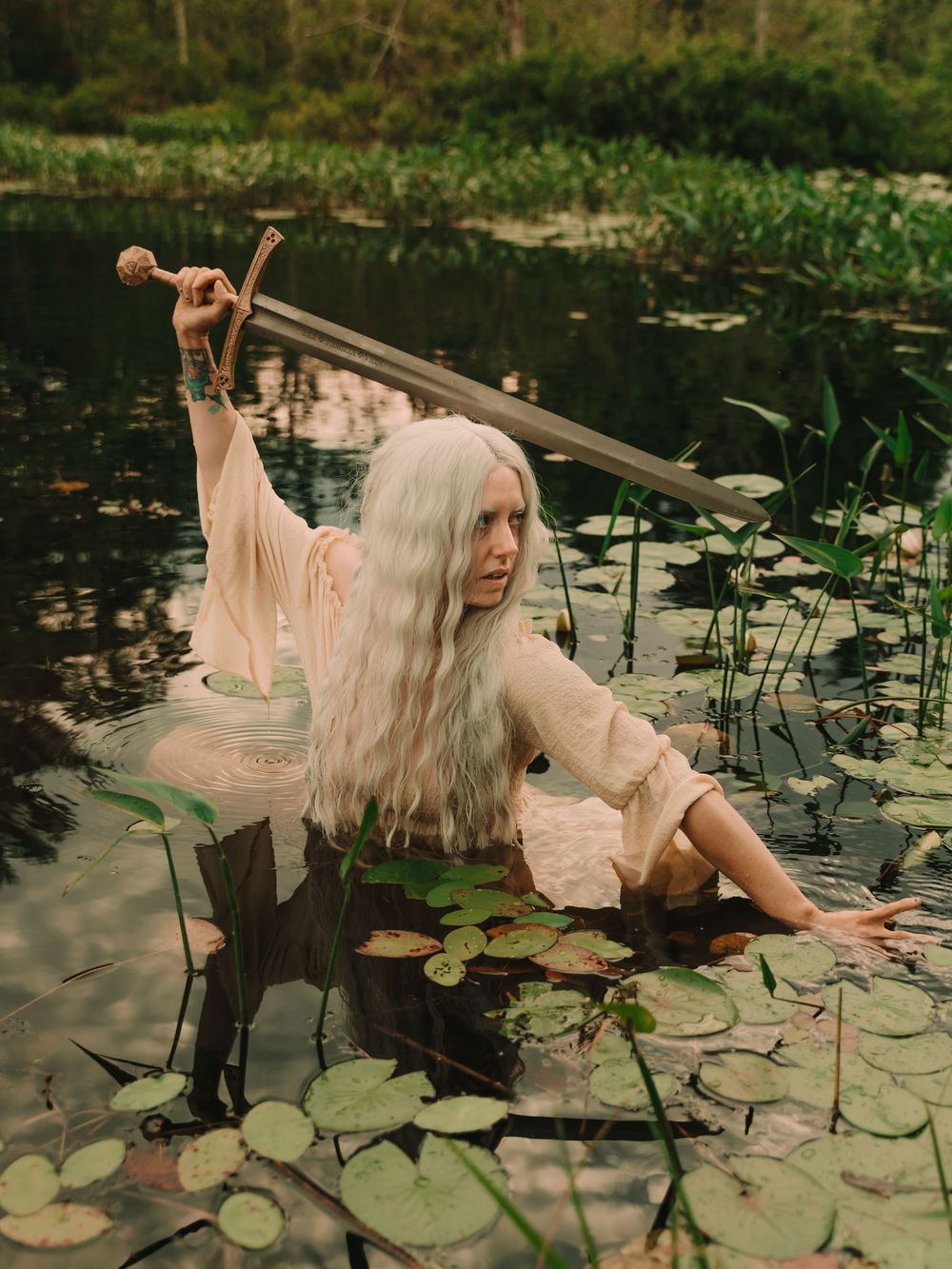 Elven fantasy woman with long white hair standing in a pond full of lilypads holding a battle sword above her head