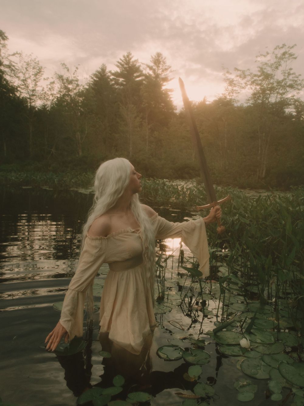 Elven fantasy woman with long white hair standing in a pond full of lilypads with a battle sword across raised to the sky