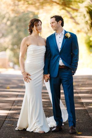 A bride in white gown and groom in navy suit hold hands while walking along a tree-lined path.