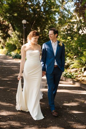 A couple in wedding attire walks together on a tree-lined path during a sunny outdoor photo session.