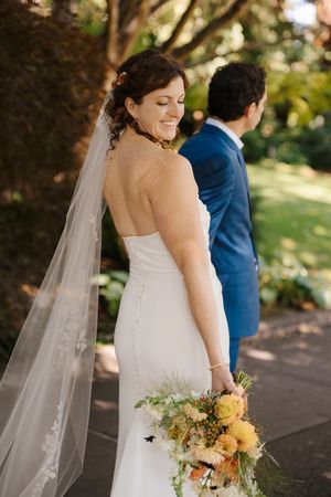 A wedding couple poses in a garden setting with the bride holding a peach and yellow bouquet while wearing a flowing veil.