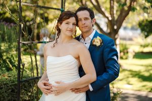 A couple in wedding attire embrace outdoors in a sunlit garden setting with lush greenery in the background.
