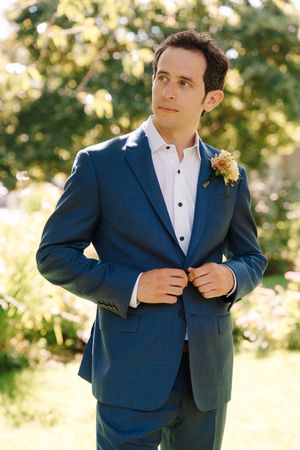 A groom in a navy suit adjusts his jacket while standing in a sunlit outdoor setting.