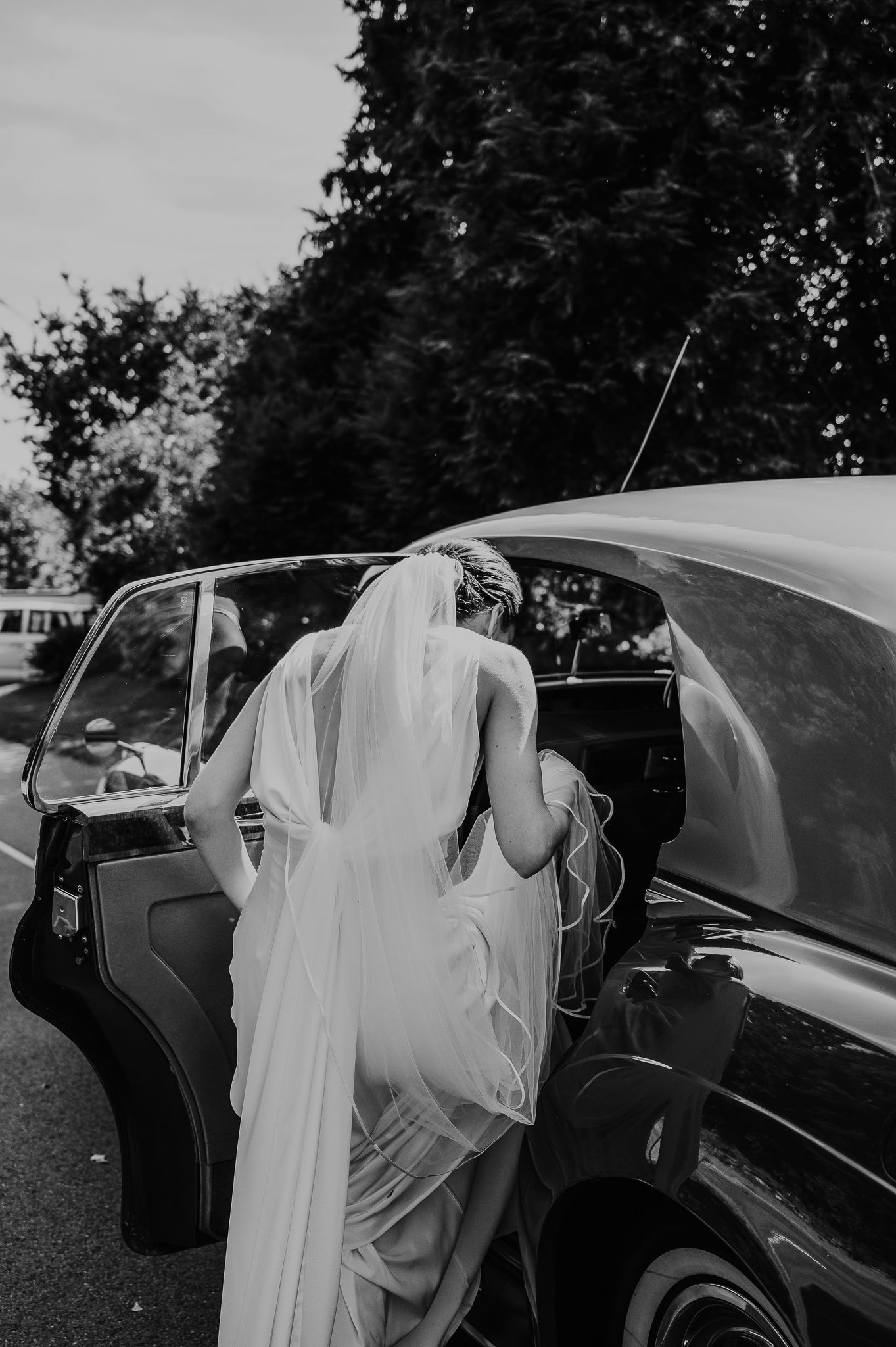 Black and white photo of bride entering vintage car with flowing veil.