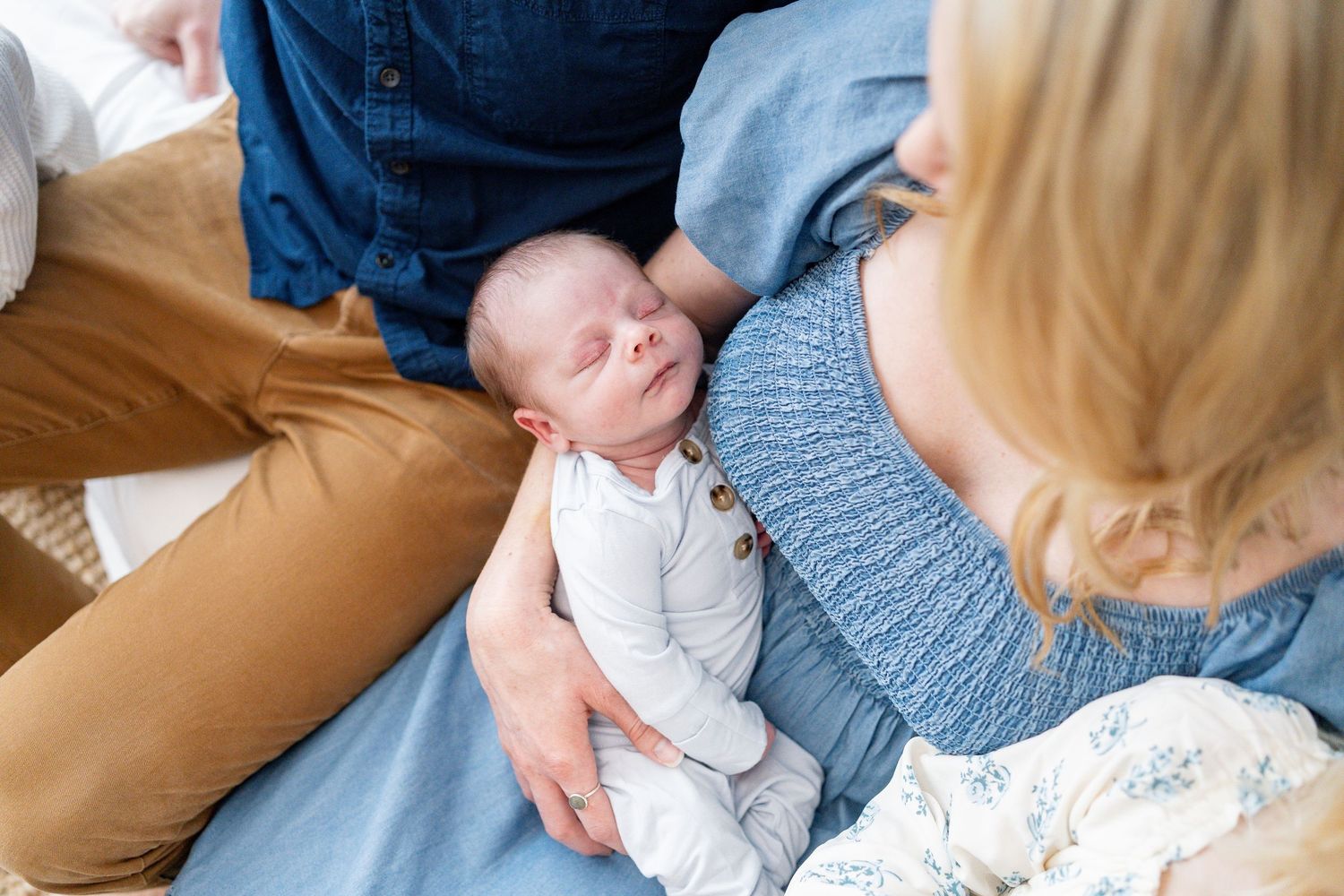A family sits together on a brown couch, sharing tender moments with a small baby in a blue onesie.