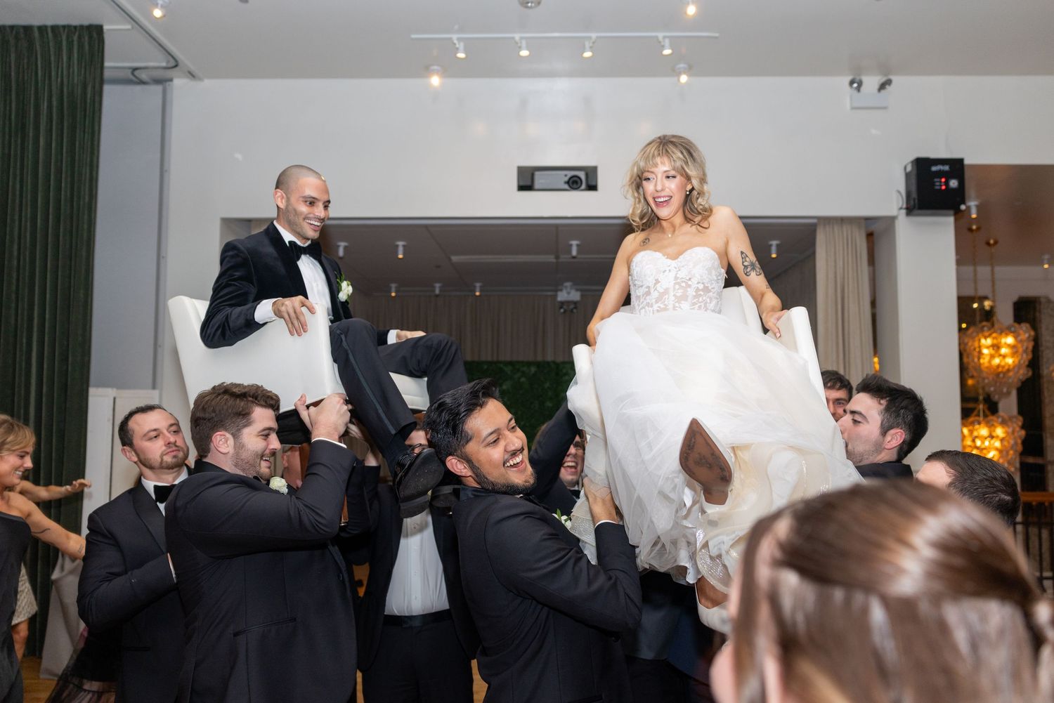 Wedding party members in formal attire pose together on elegant wooden benches at an indoor reception venue.