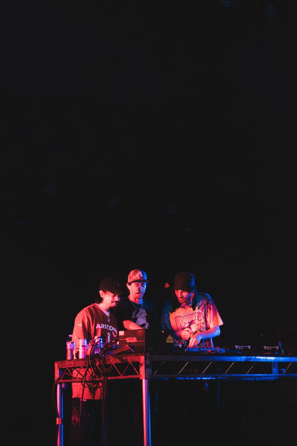 Musicians perform on stage with electronic equipment under dramatic red and blue lighting against a dark background.