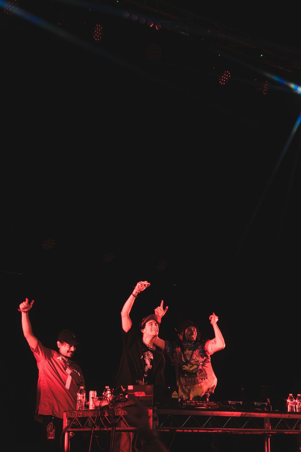 Performers raise their arms in celebration behind equipment tables during a dark concert lit with red lights.