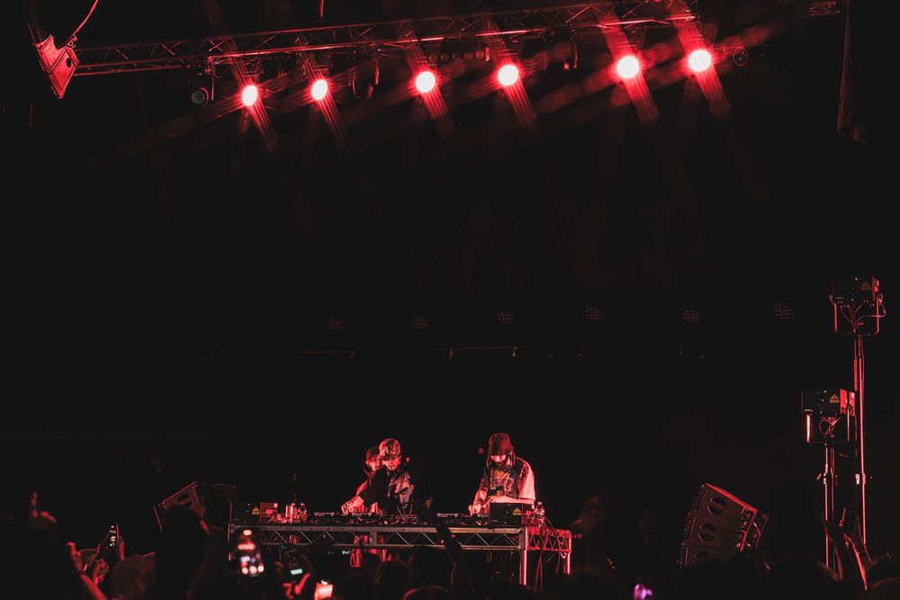 Red stage lights illuminate performers at DJ booth during dark nighttime music performance.