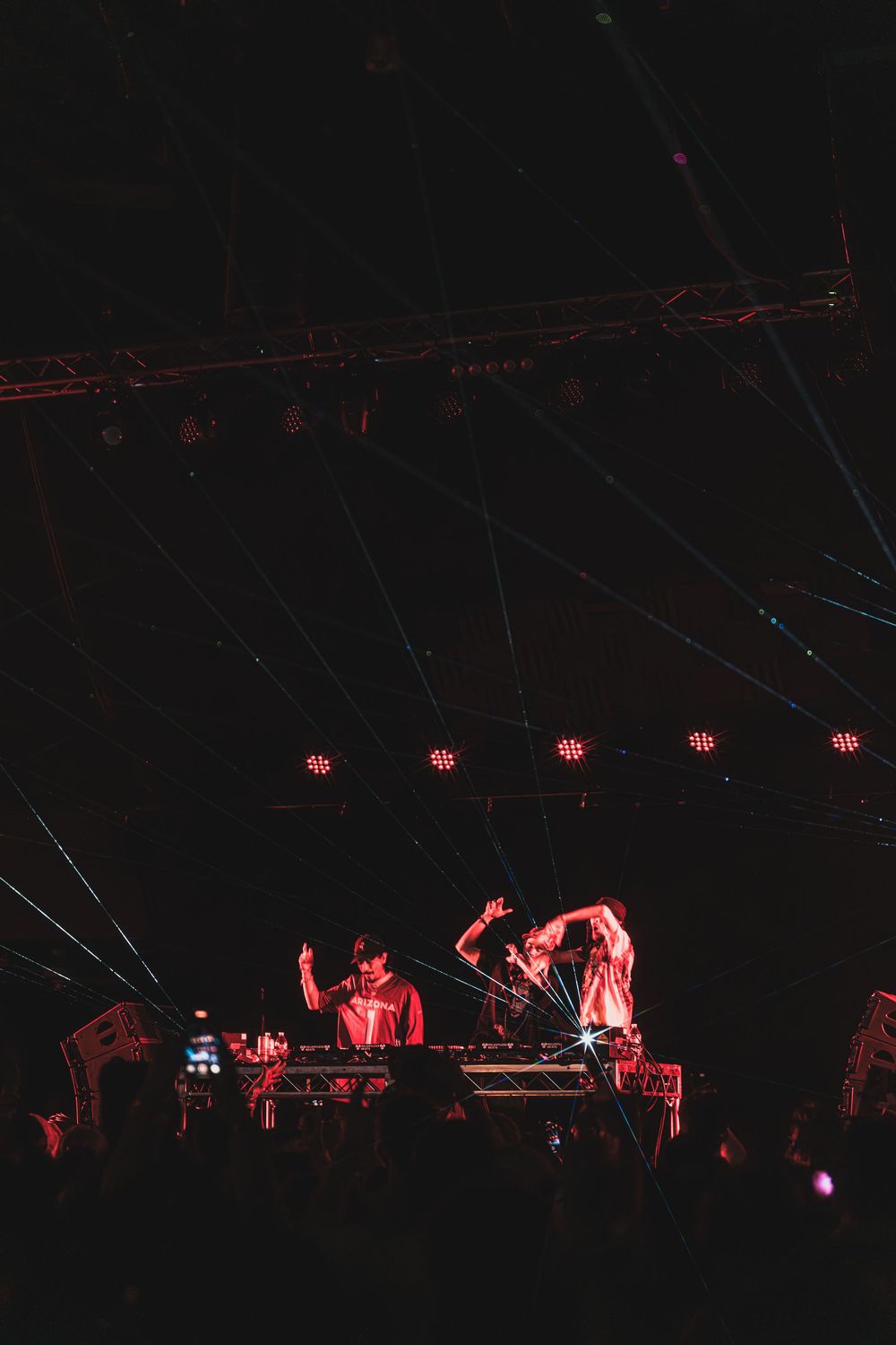 DJs perform on stage under dramatic red lighting at a nightclub with overhead rigging visible in the dark atmosphere.