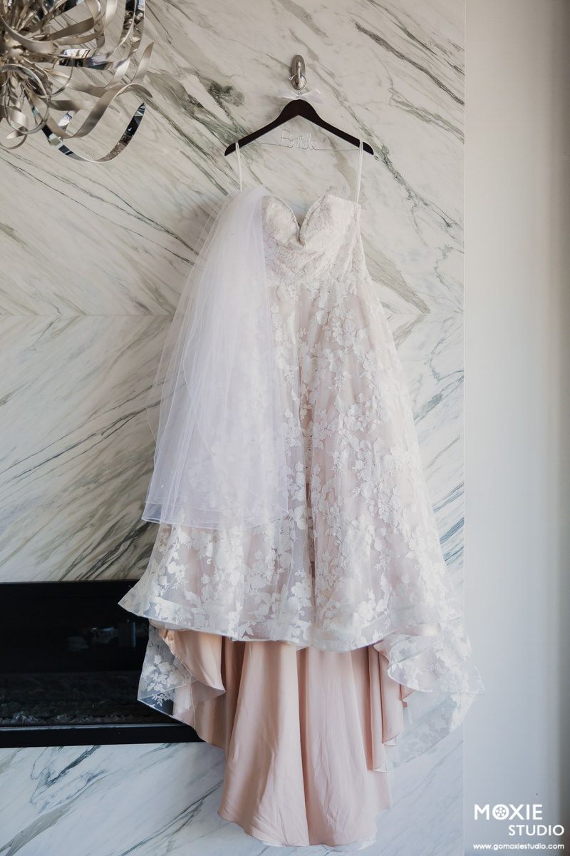 A delicate blush pink and white lace wedding dress hangs elegantly against a marble-patterned wall.