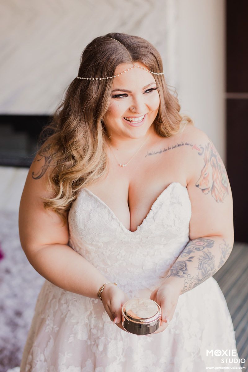 A bride in a white lace wedding dress with tattoos holds a beverage while posing for bridal portraits.