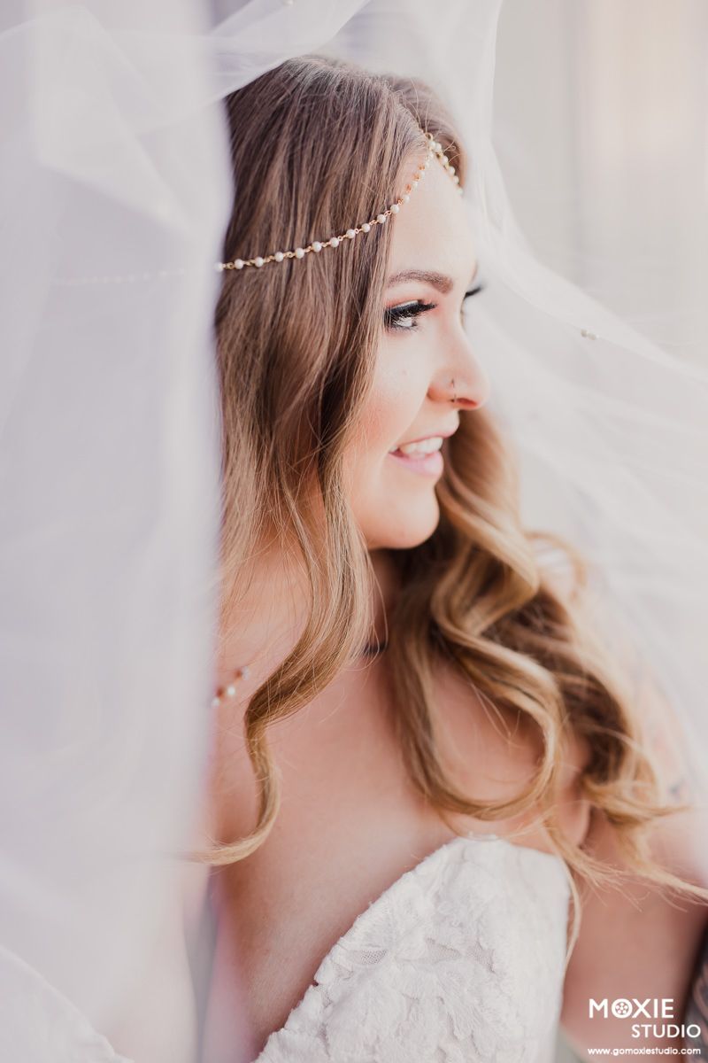 Bride in white lace dress with delicate pearl headpiece and flowing veil smiles in soft natural light.