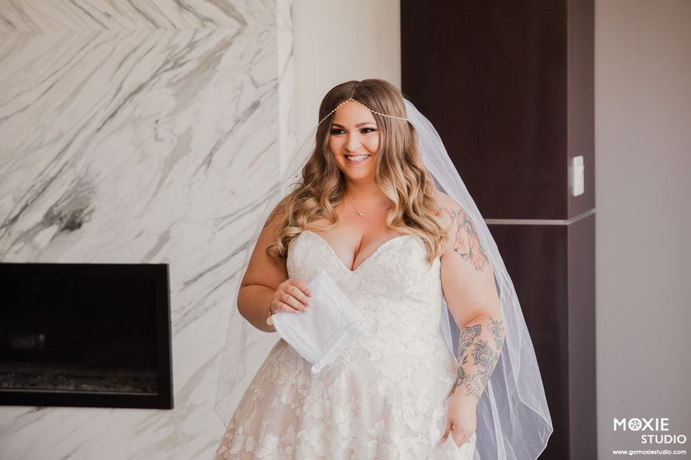 A bride in a white lace wedding gown stands in an elegant marble-walled room preparing for her special day.