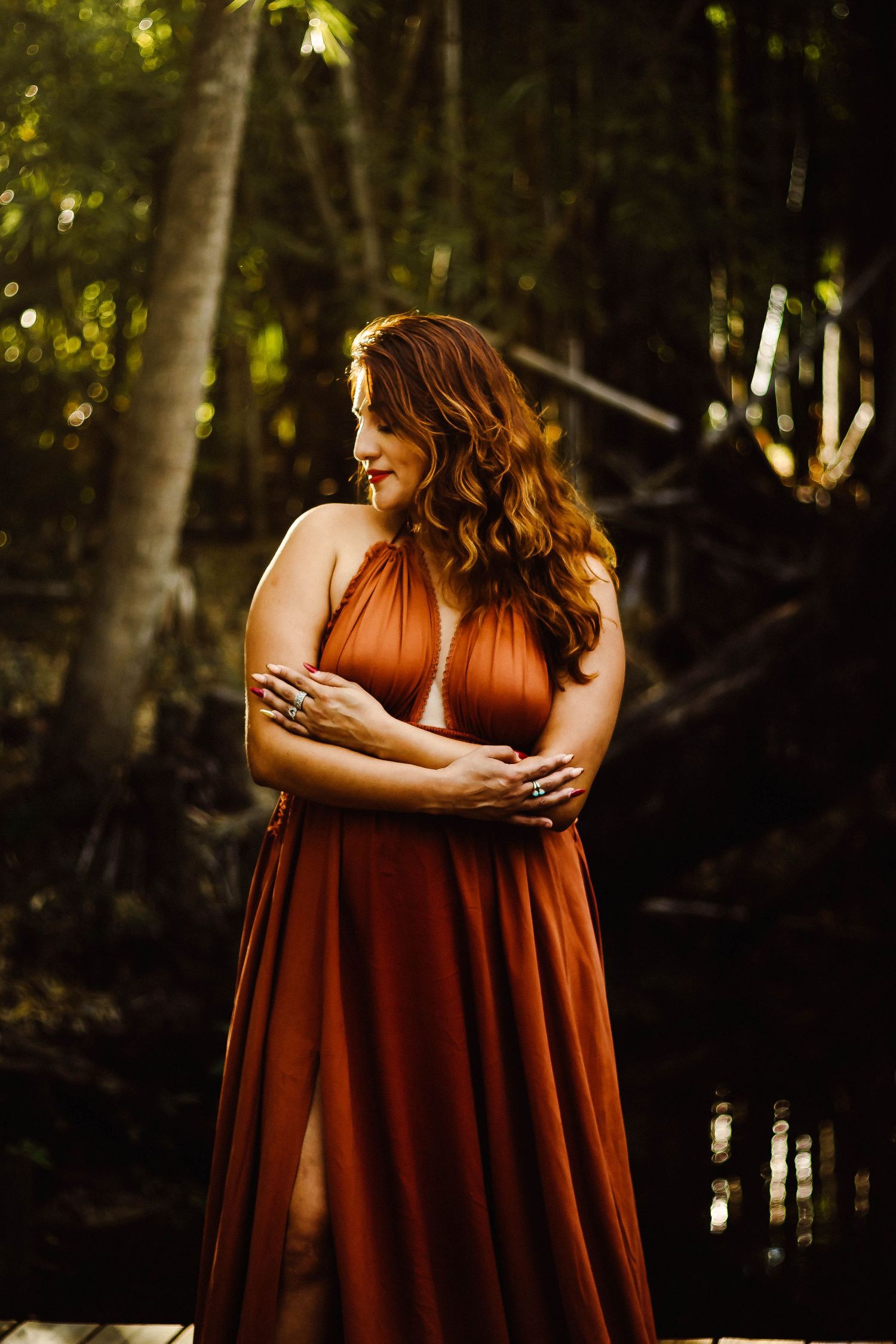 Two figures in rust-colored dresses embrace during golden hour in a forest setting.