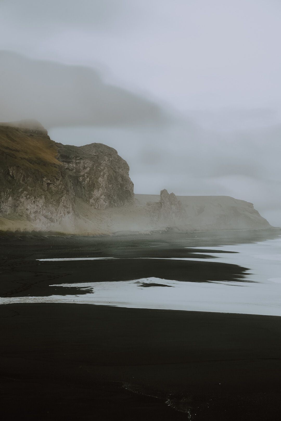 Dramatic black sand beach with misty cliffs and rocky coastline along Iceland's southern shore in moody weather.