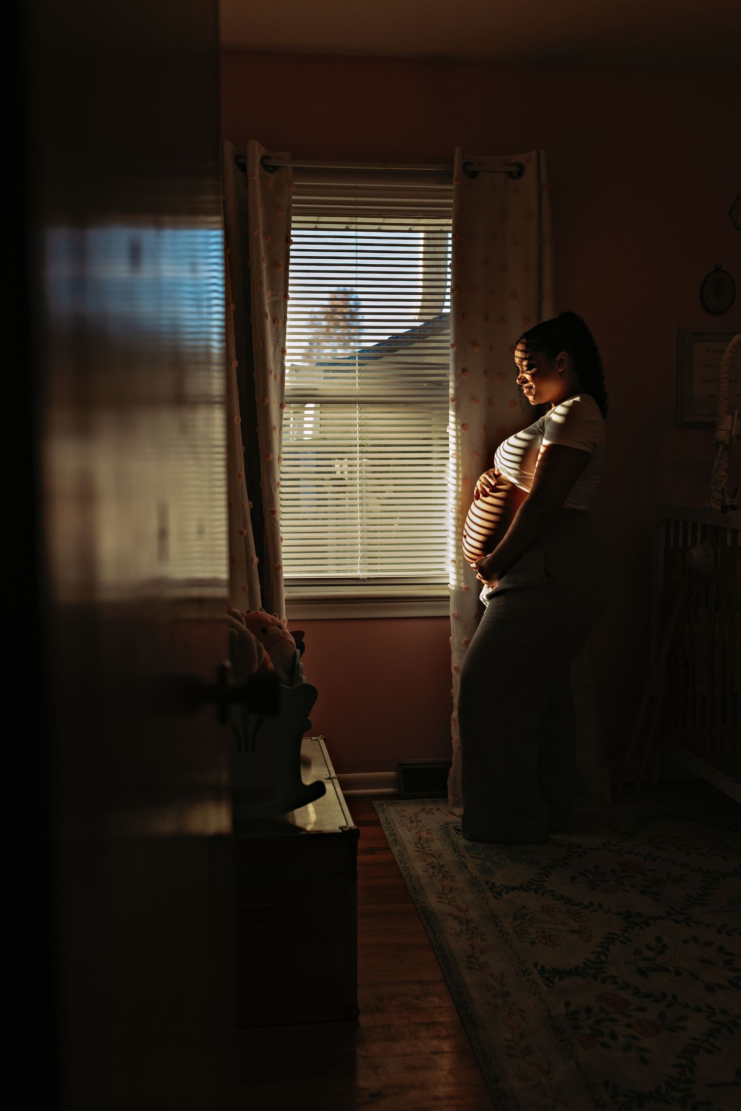 Silhouette of person standing by window with sunlight streaming through blinds in dark moody room.