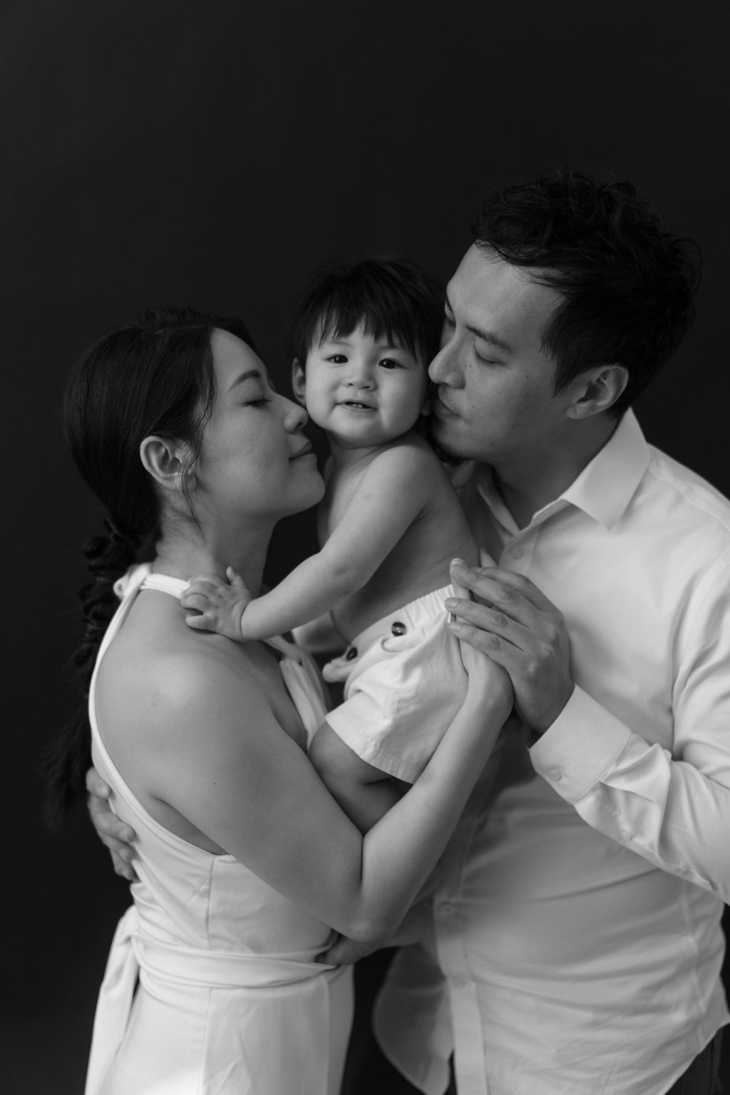 A tender black and white family portrait showing parents kissing their smiling toddler against a dark background.