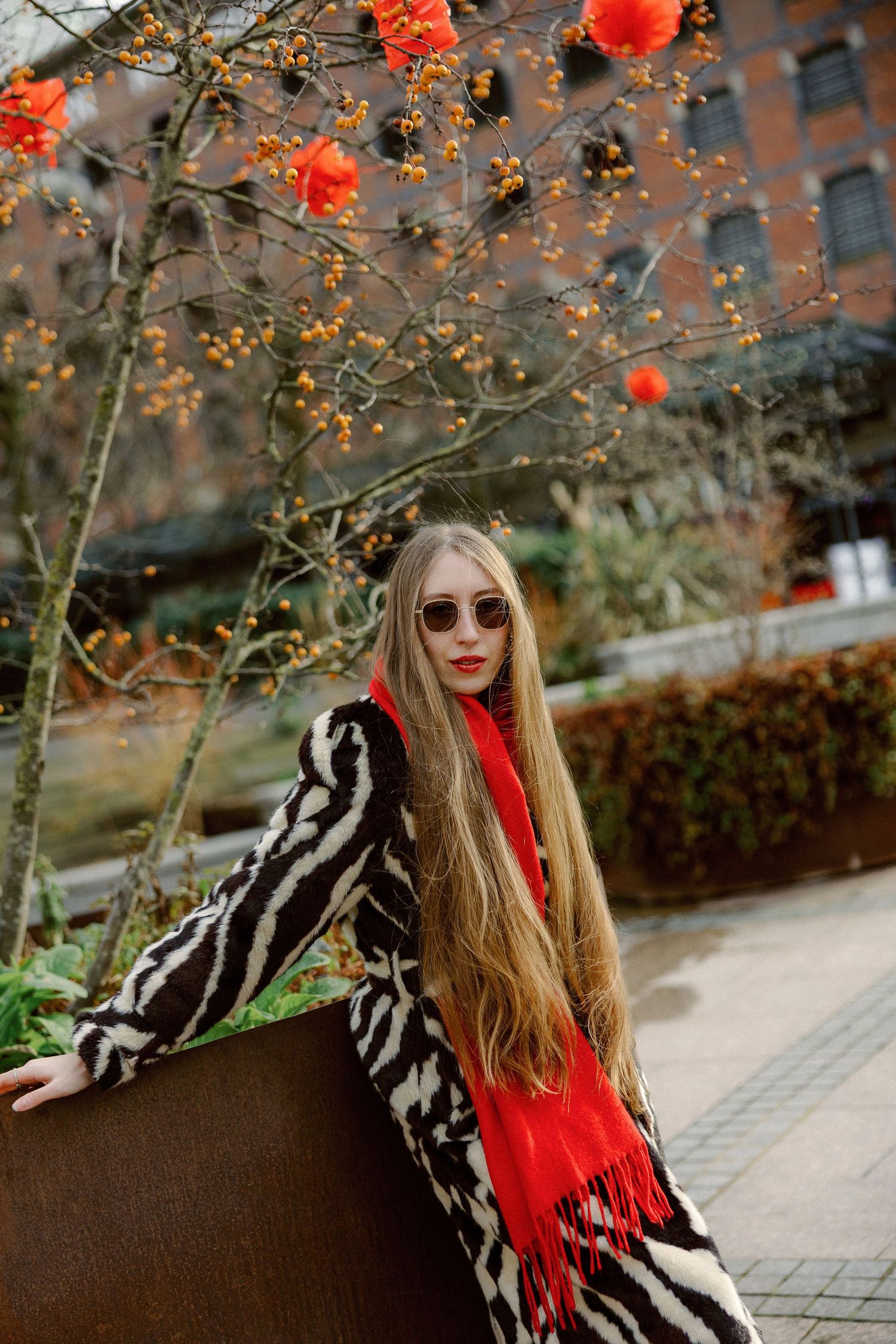Stylish winter fashion with zebra print coat and bright red scarf under blooming orange flowers in urban setting.