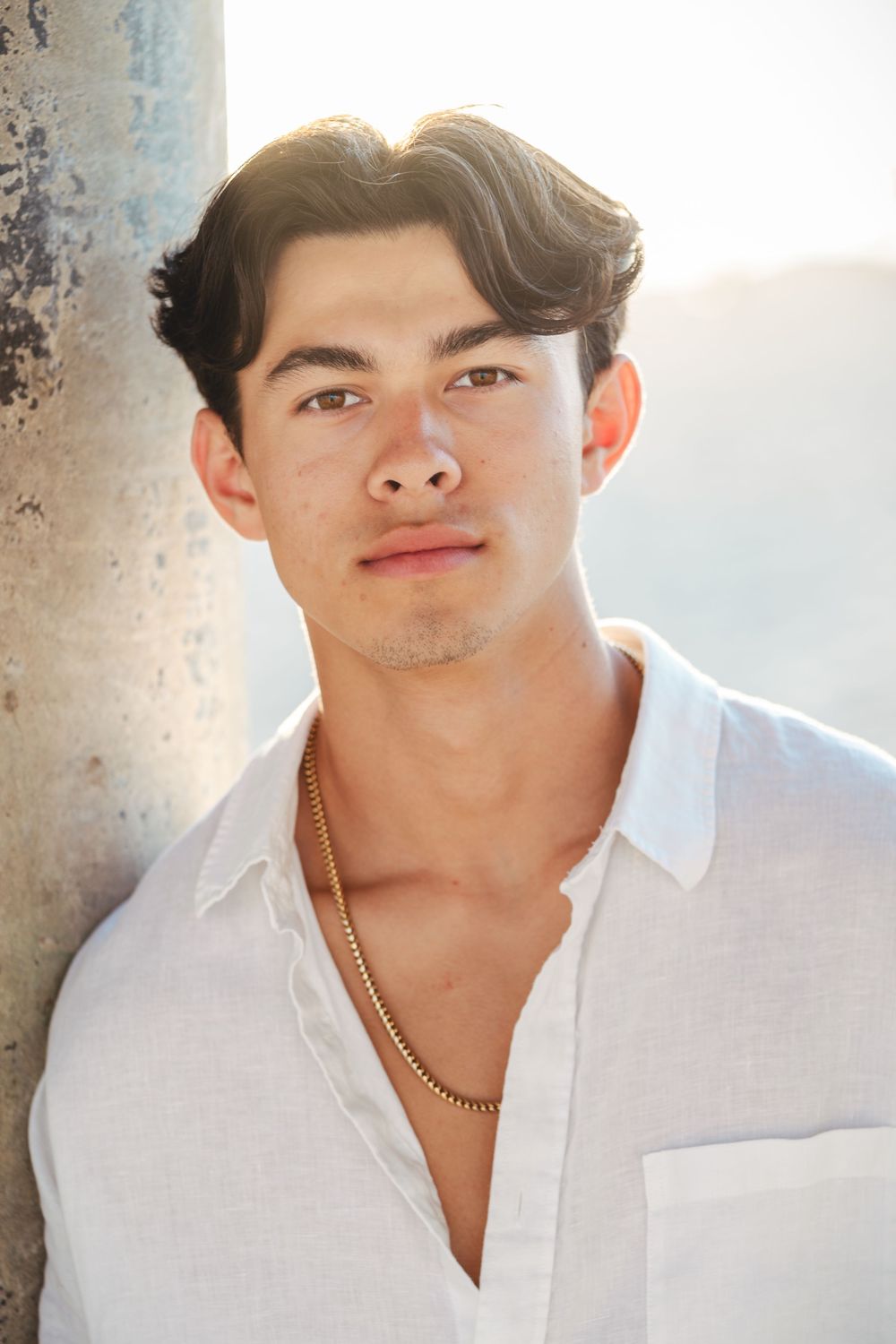 Portrait of a young Asian man in a white button-down shirt with a gold chain necklace in warm sunlight.