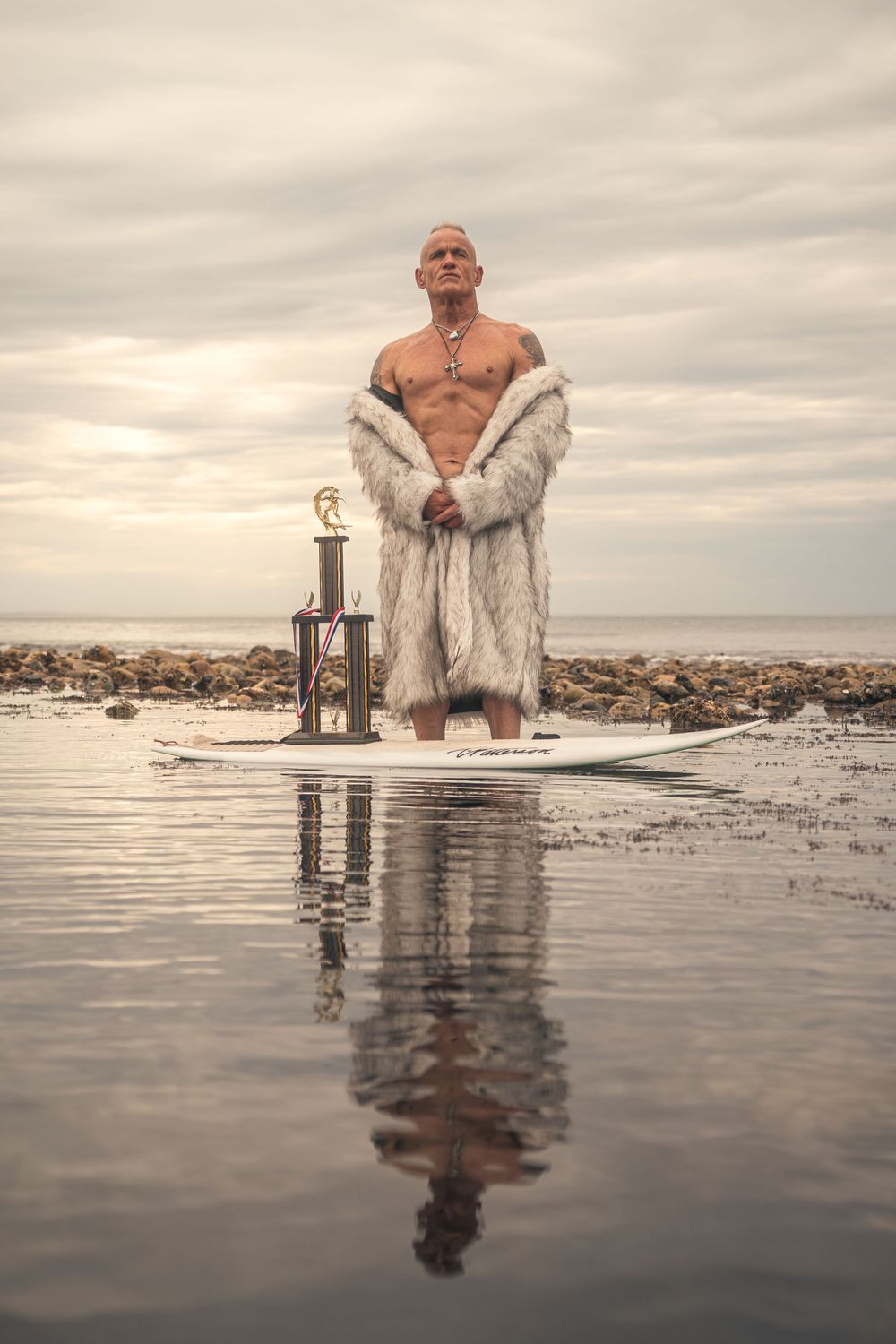 A person wearing a white fur coat sits on a chair in shallow water with reflections at sunset.