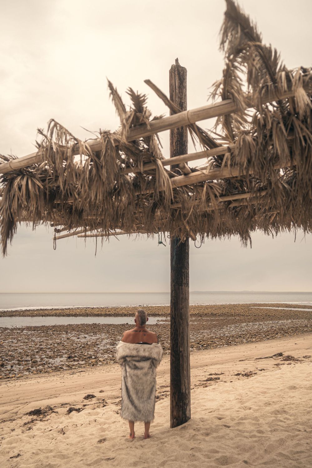 A rustic beach shelter with thatched palm fronds provides shade along a sandy shoreline on a cloudy day.