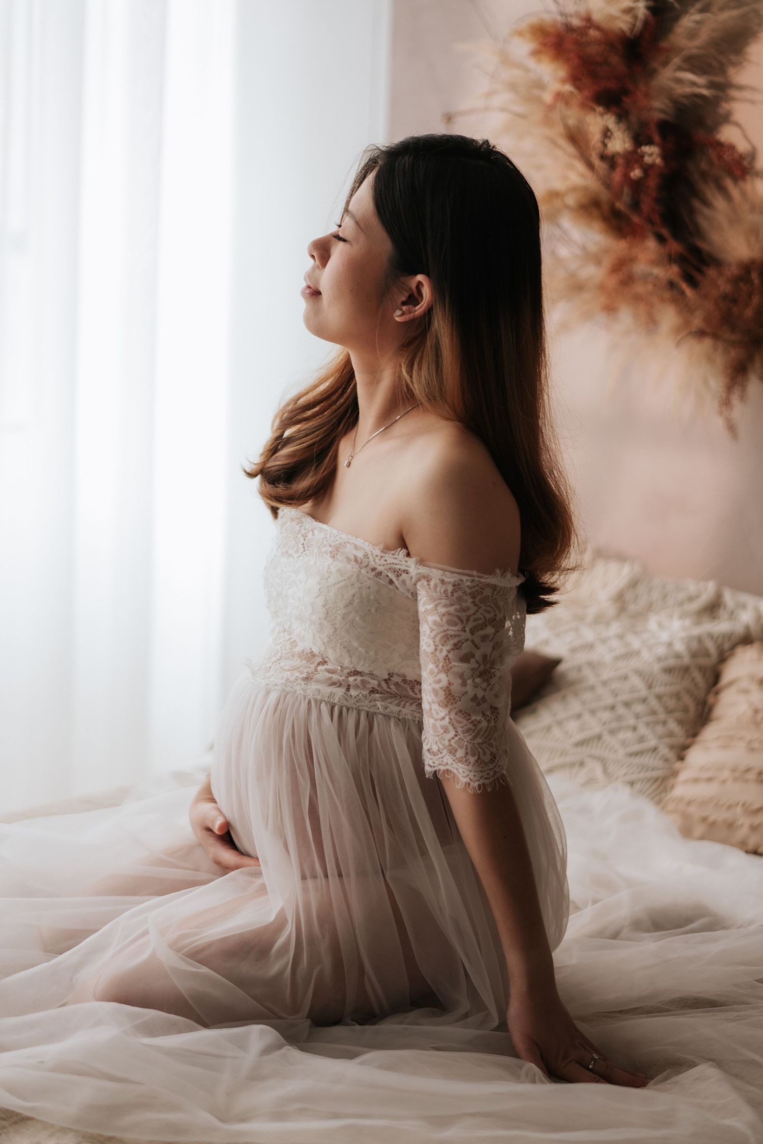 Ethereal maternity portrait of woman in flowing white lace dress sitting on bed with soft natural lighting.