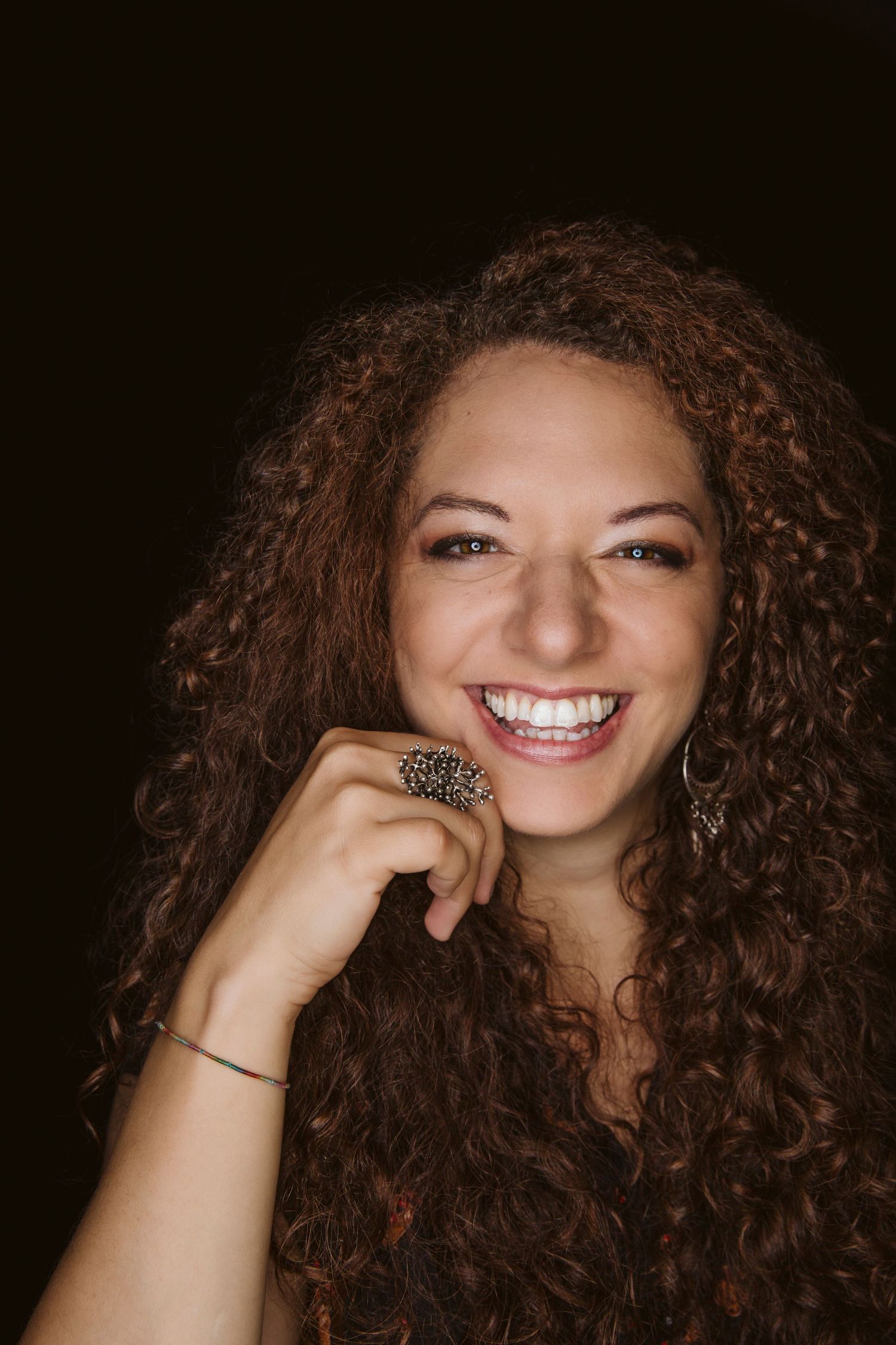 Portrait of a woman with natural curly brown hair and a bright smile wearing a statement ring against a dark background.