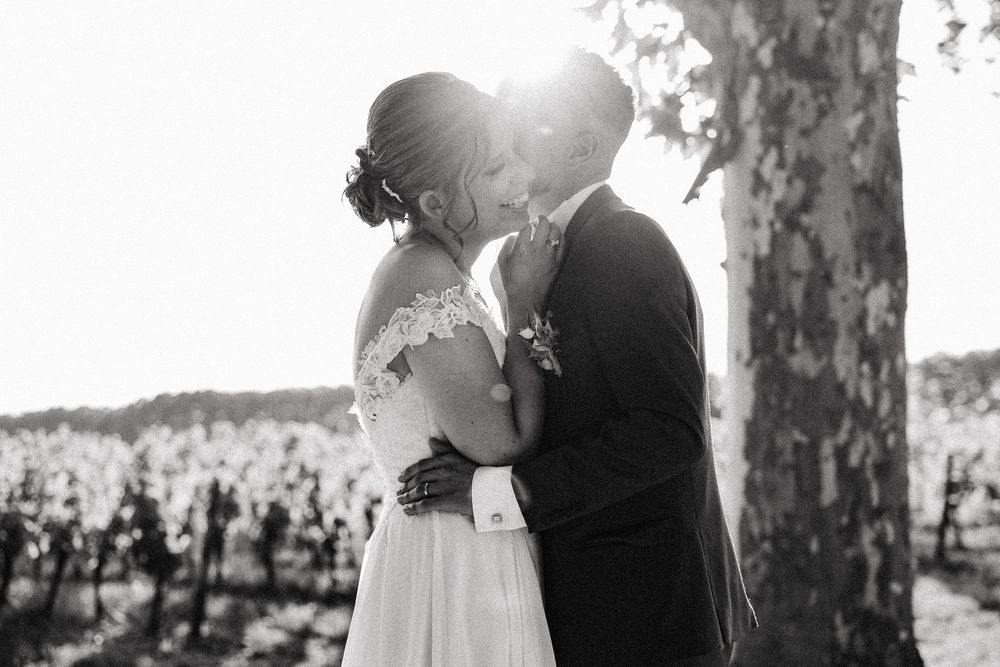 Black and white photo of bride and groom kissing passionately in front of a sun-drenched vineyard.