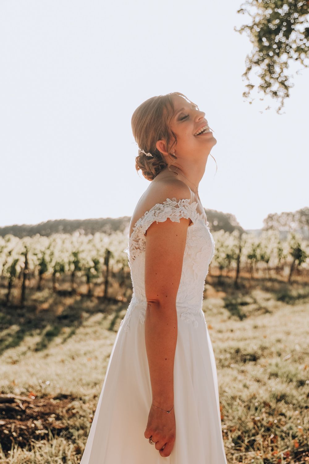 Radiant bride in an off-shoulder lace gown laughing joyfully in golden sunlight beside a vineyard.