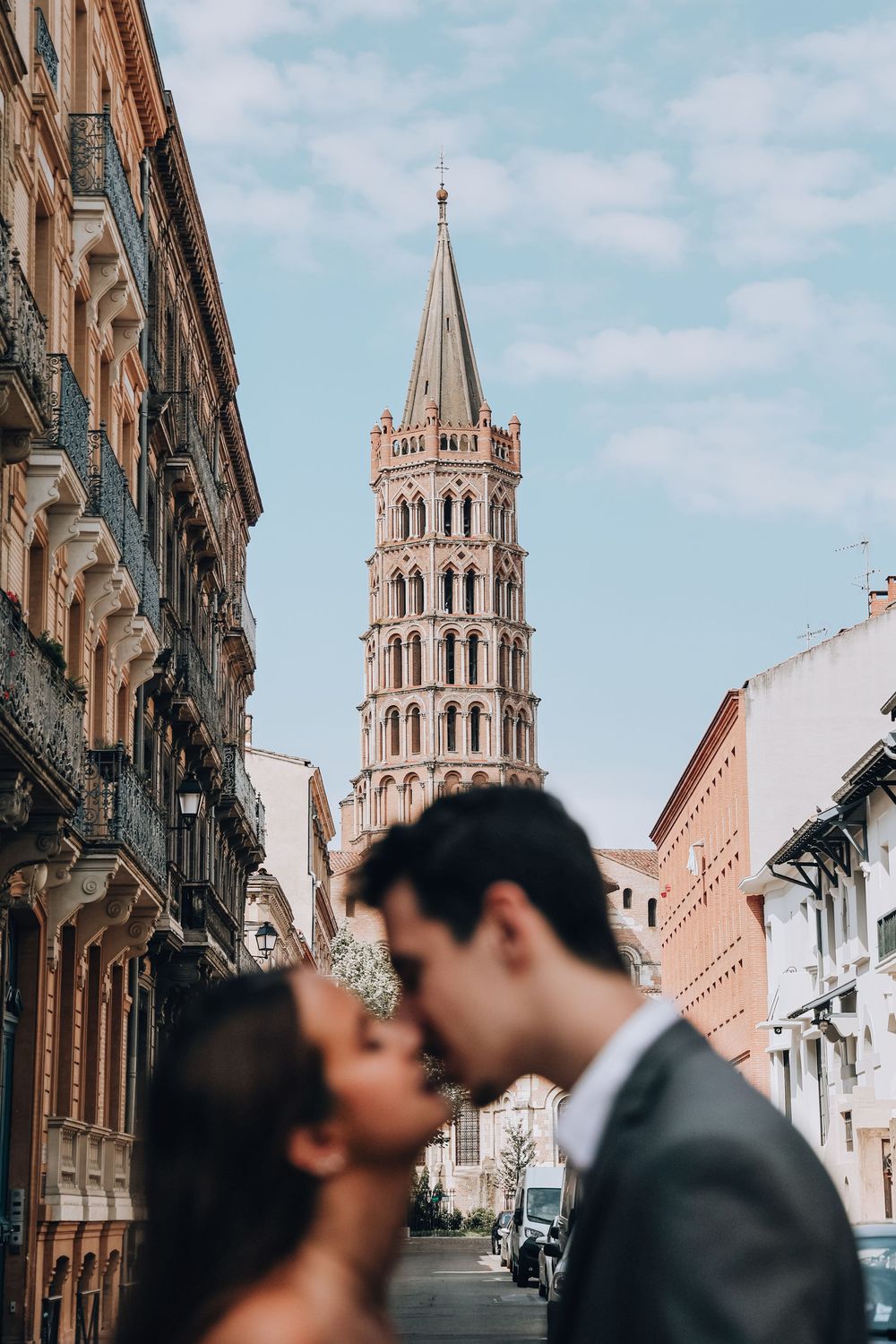 Couple sharing a kiss on a street in Toulouse with the Basilica of Saint-Sernin tower in the background.