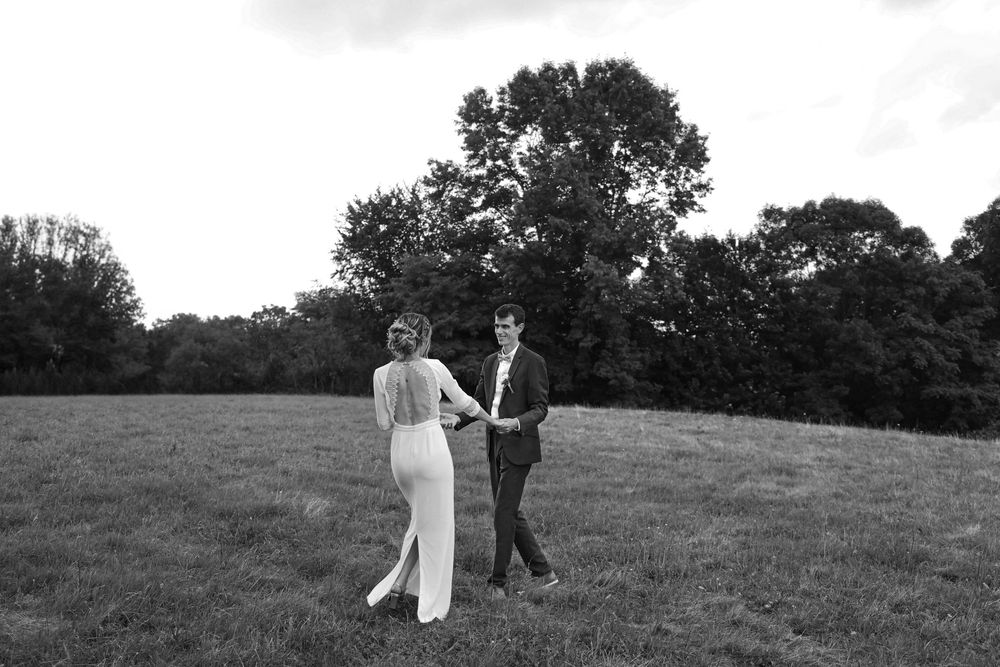 Black and white photo of bride and groom walking hand in hand through a lush green countryside field.