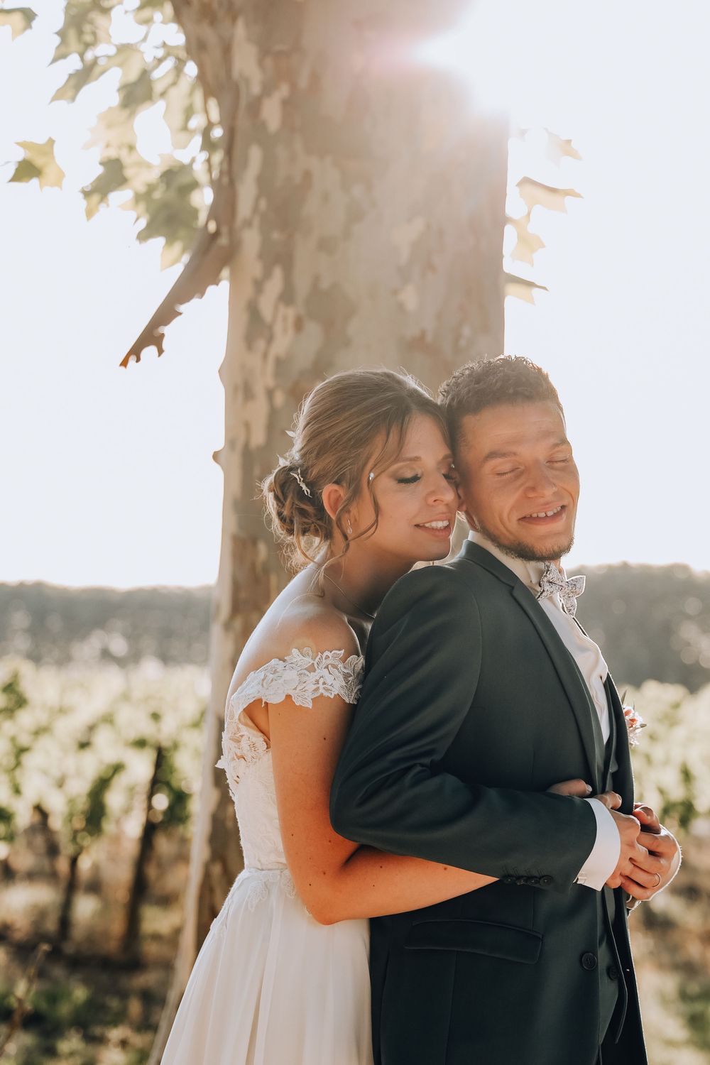 Bride hugging groom from behind near a sunlit tree with vineyard rows in the golden background.