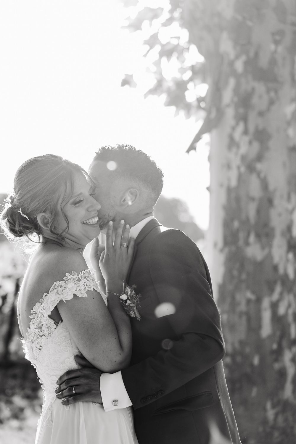 Black and white photo of groom tenderly kissing brides cheek in bright sunlight near a tree.