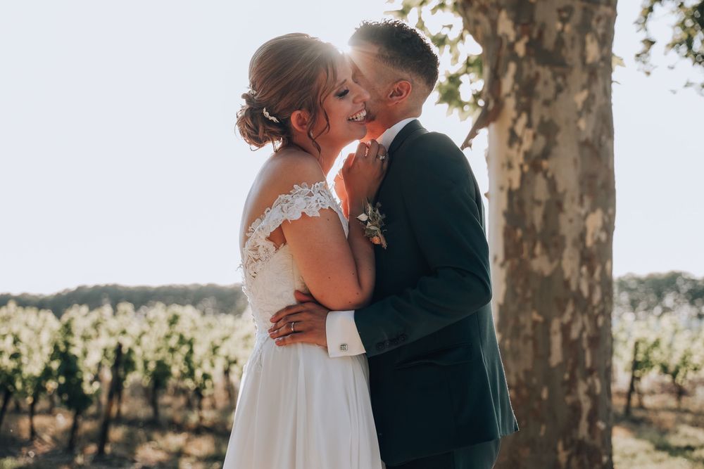 Bride and groom sharing a romantic kiss in front of a sunlit vineyard with a tree beside them.