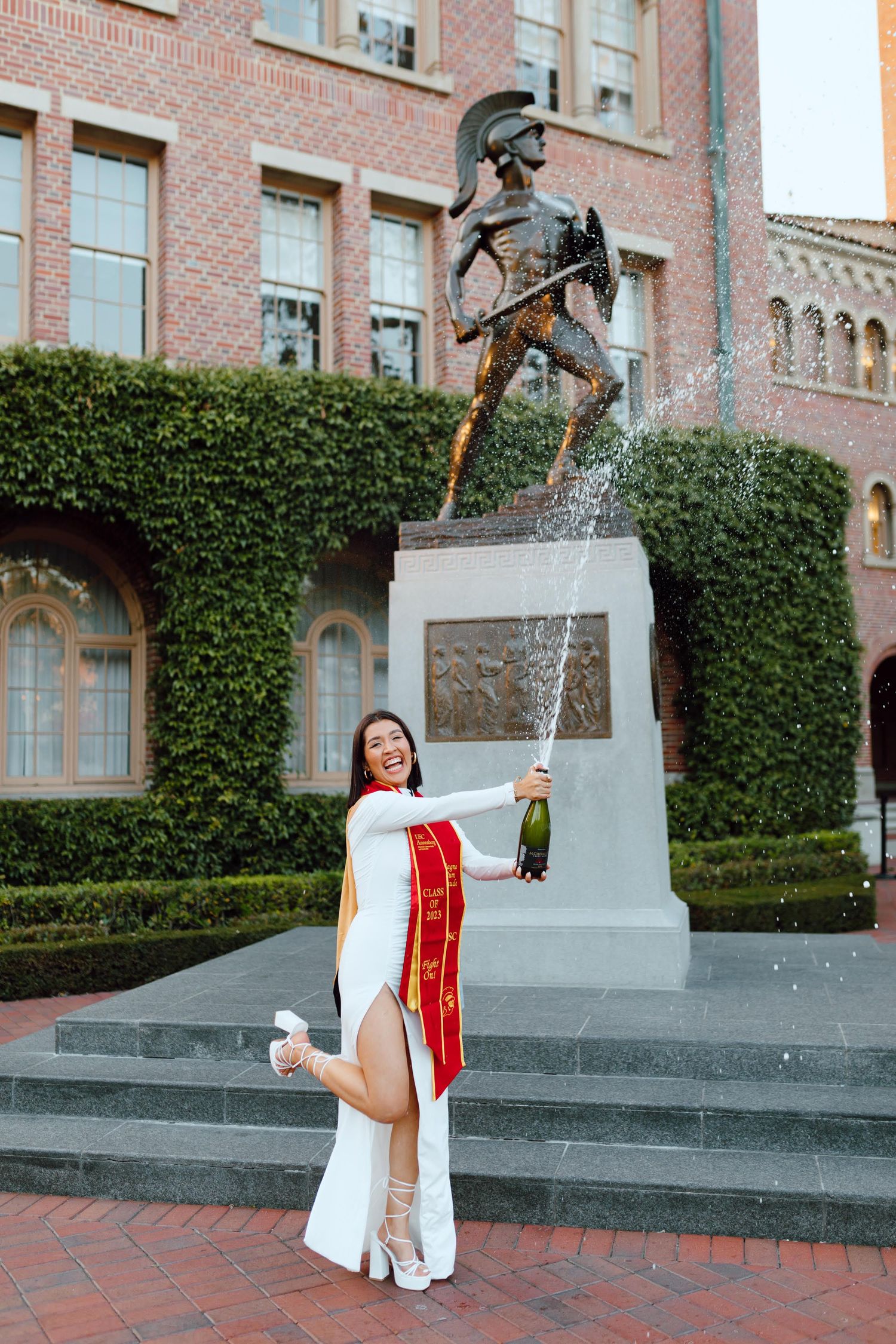 Graduation celebration photo in front of the Tommy Trojan statue at USC campus with ivy-covered brick building backdrop.