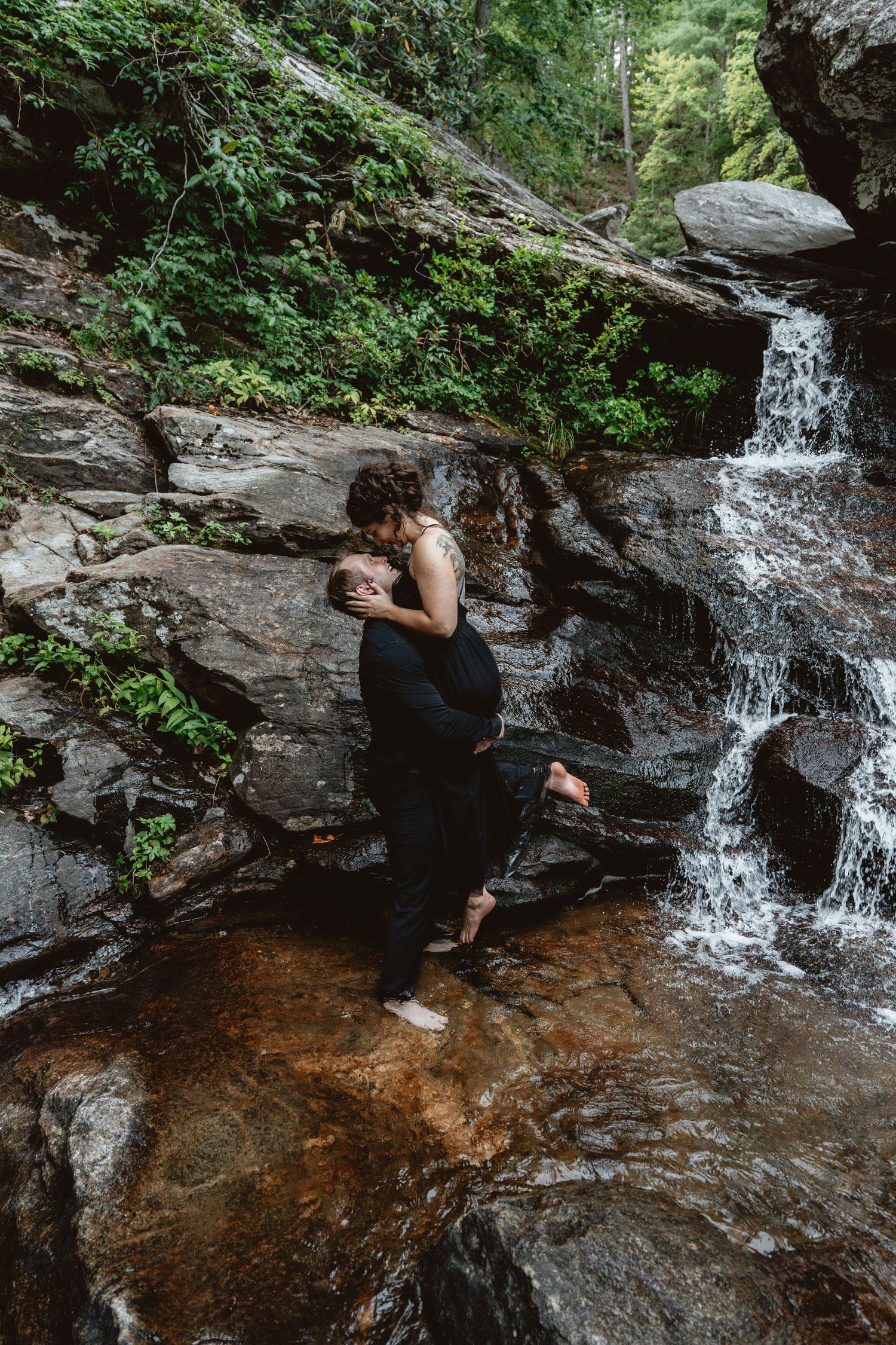 A person takes photos near a cascading waterfall in a lush forest setting with rocky outcrops and a shallow pool.