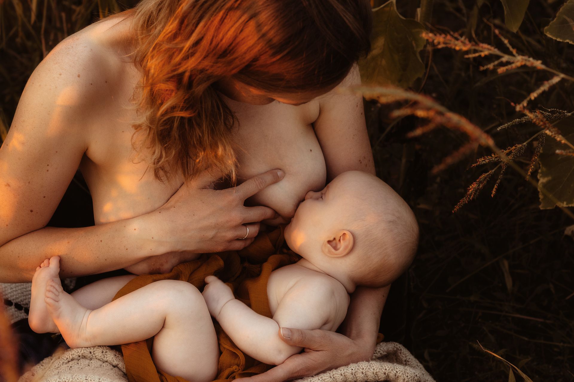 A tender moment between a mother and baby nursing in nature with warm sunlight filtering through trees.