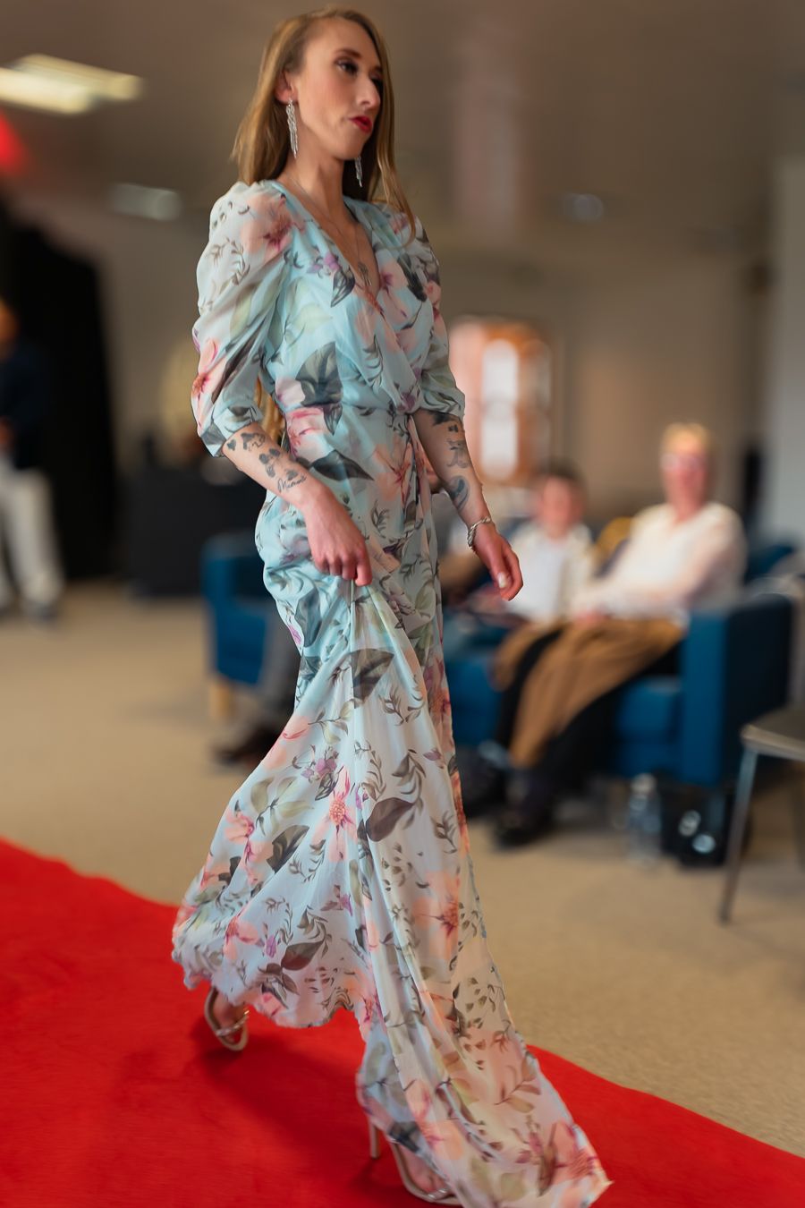 Model walks the runway in a flowing light blue floral gown with puff sleeves on red carpet at fashion event.