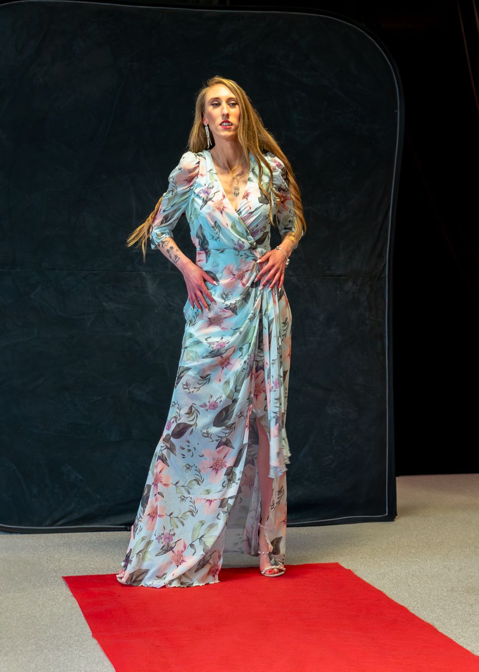A model poses in a flowing floral blue and white gown on a red carpet against a dark backdrop.