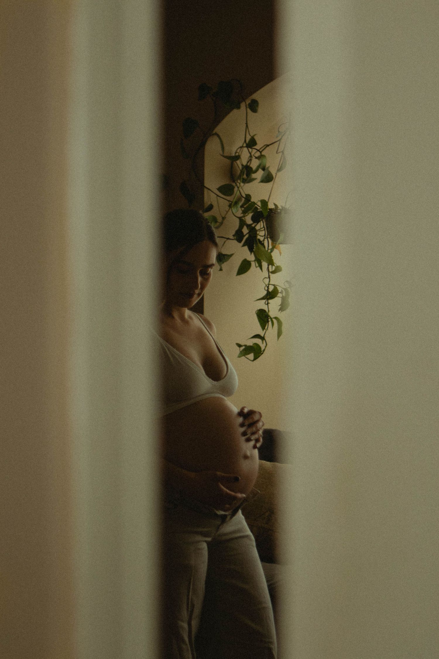 Pregnant woman cradling baby bump standing near indoor plants viewed through curtains.