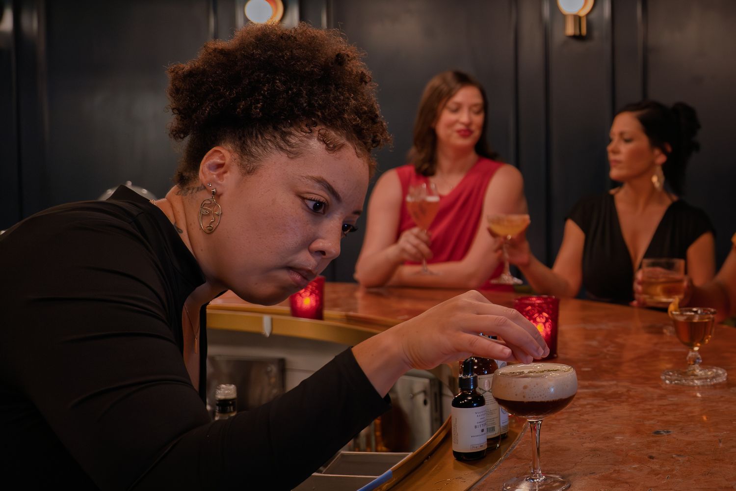 A bartender carefully places a garnish on a freshly made cocktail while two well dressed women enjoy a drink and conversation at the bar