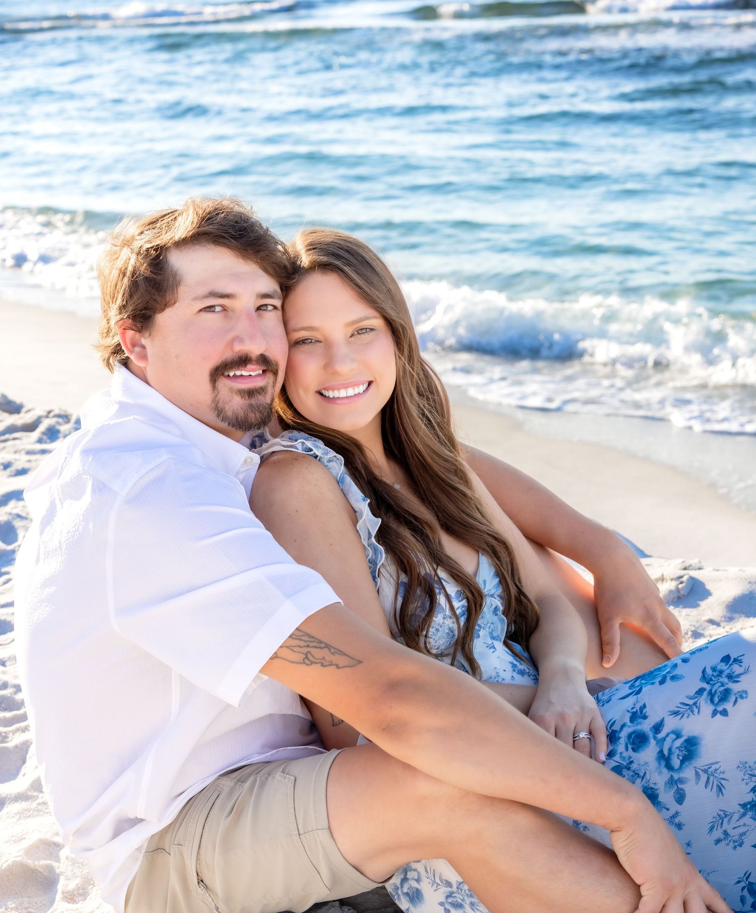 A couple embraces on a sunny beach with waves in the background wearing white and blue coordinated outfits.