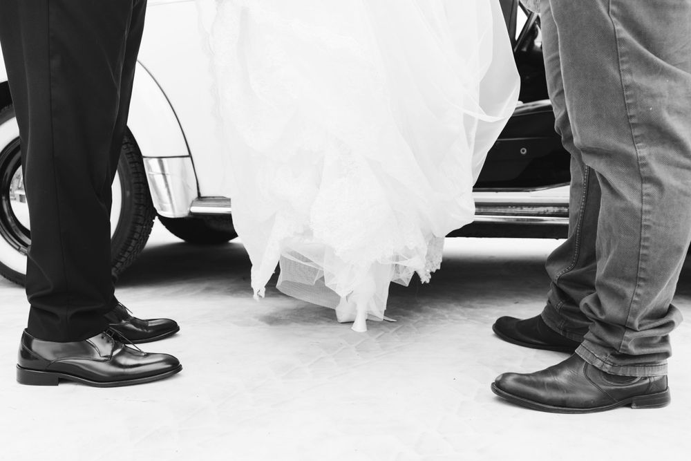Colpo di dettaglio in bianco e nero delle scarpe da sposa e dell'orlo del vestito tra due paia di scarpe formali da uomo, fratello e papà.