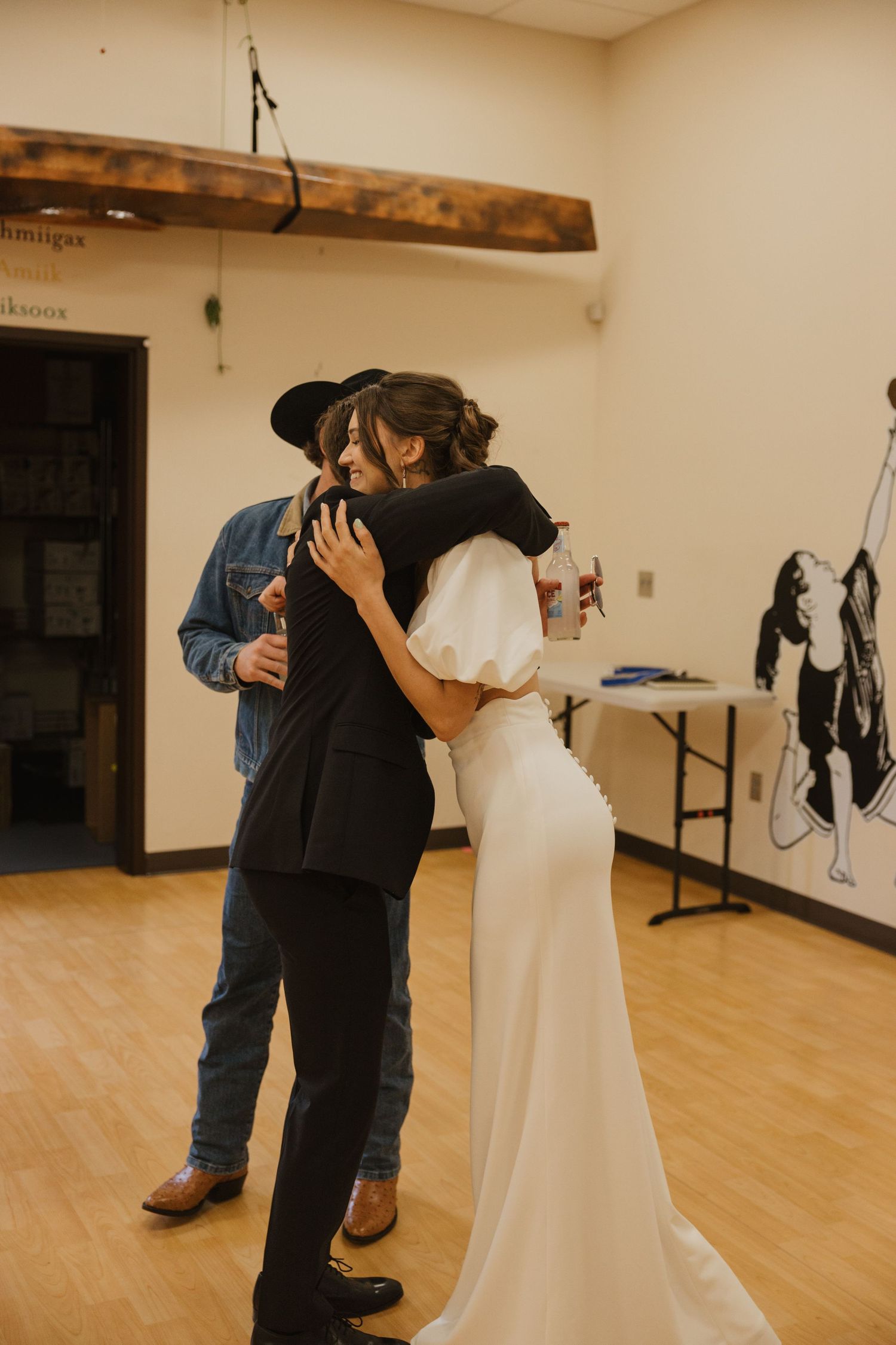 Bride in white gown embracing a guest in black outfit at a wedding venue.