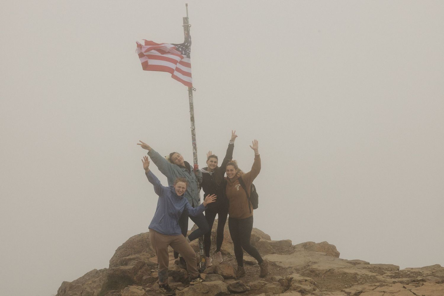 Four people celebrating at a mountain summit next to an American flag in foggy misty weather conditions.