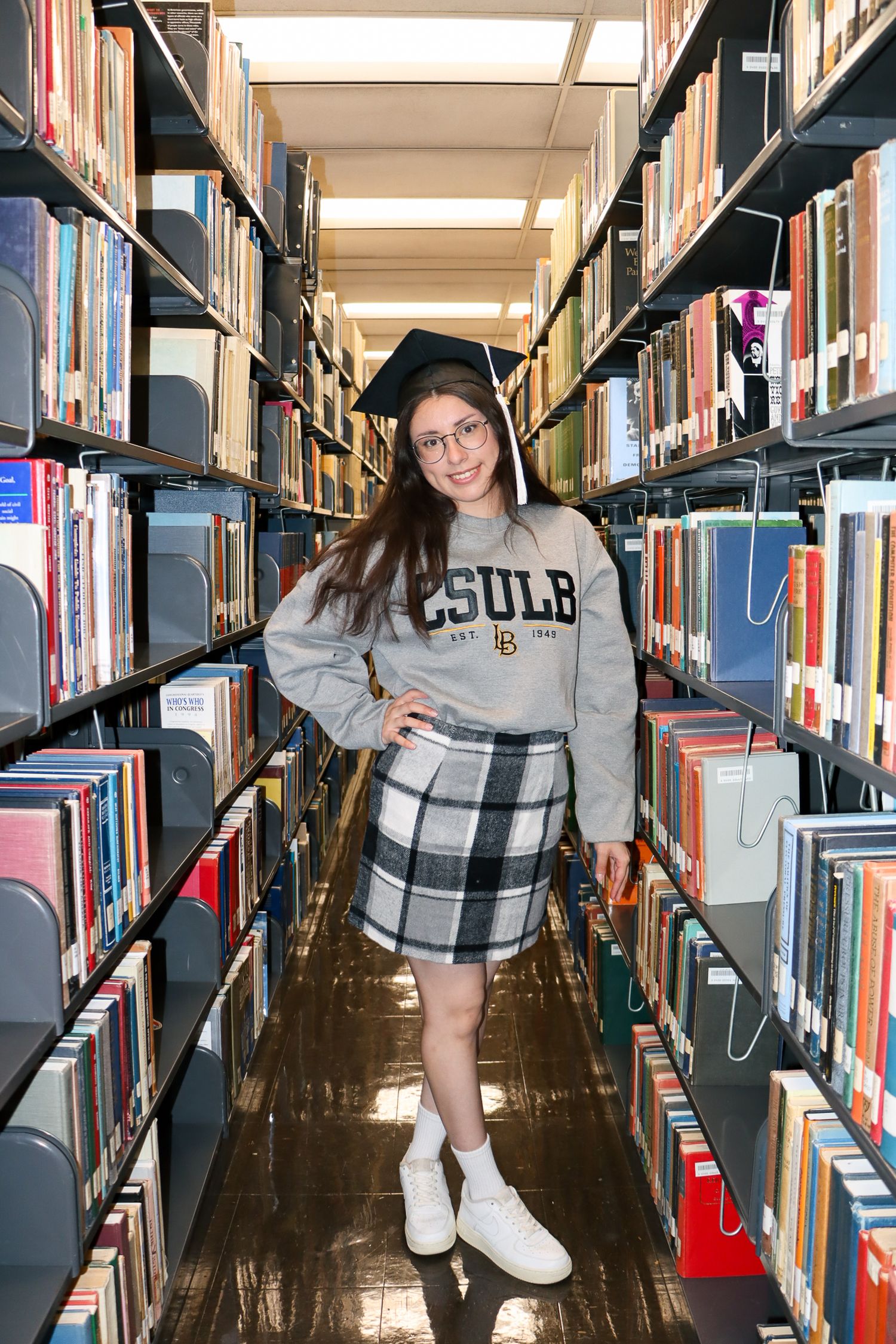 A person wearing a gray sweater and sneakers browses books in a library aisle between tall shelving units.