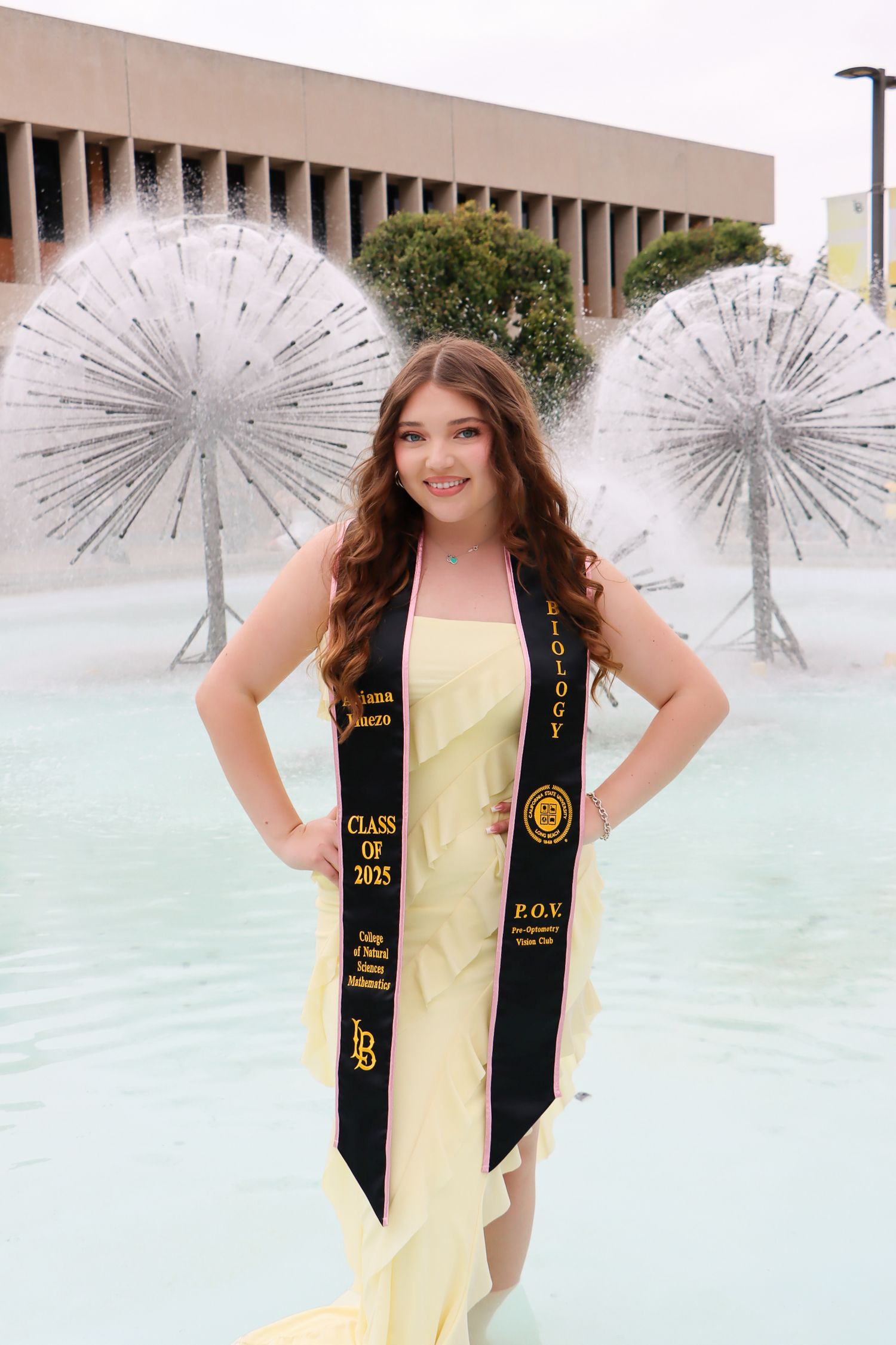 Graduate wearing yellow dress and black sash poses with decorative silver umbrellas by fountain during graduation photoshoot.