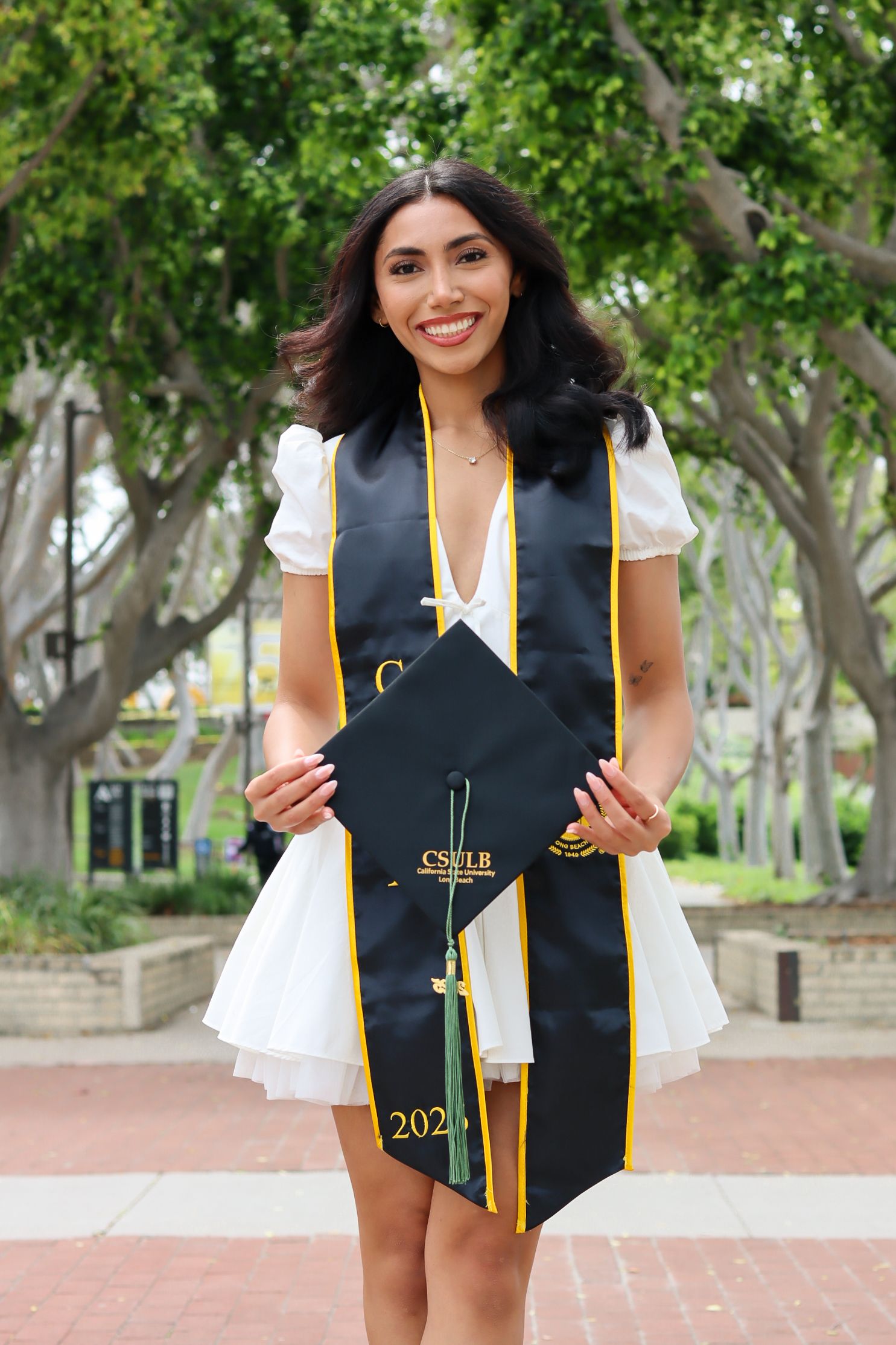 A graduate in a black graduation stole and white dress poses outdoors against a backdrop of green trees.